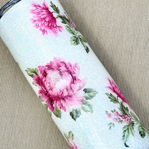 May include: A light blue glitter tumbler decorated with pink peony floral designs and green leaves. The stainless steel tumbler has a black lid. The floral pattern is a watercolor style. The tumbler is designed to keep drinks cold or hot.