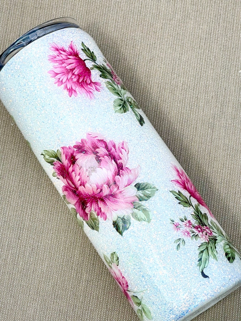 May include: A light blue glitter tumbler decorated with pink peony floral designs and green leaves. The stainless steel tumbler has a black lid. The floral pattern is a watercolor style. The tumbler is designed to keep drinks cold or hot.