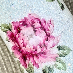 May include: Close-up of a tumbler with a light blue glitter background. The tumbler features a large floral design with pink and white petals and green leaves. The design is accented with glitter, creating a sparkling effect.