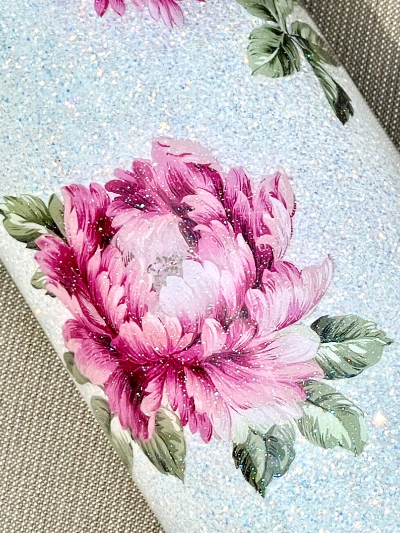 May include: Close-up of a tumbler with a light blue glitter background. The tumbler features a large floral design with pink and white petals and green leaves. The design is accented with glitter, creating a sparkling effect.