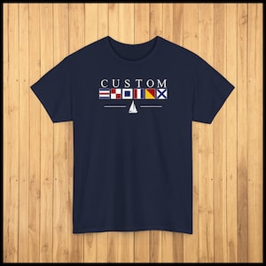 May include: Navy blue t-shirt with the word "CUSTOM" above a row of nautical signal flags and a white sailboat graphic. The design is centered on the front of the shirt.