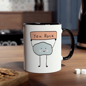 May include: White ceramic mug with a black rim and handle. The mug has a cartoon rock holding a sign that says "You Rock".