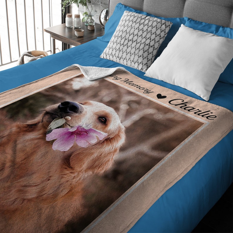 May include: A beige fleece throw blanket with a photo of a golden retriever holding a pink flower. The blanket has a brown border and the text "Loving Memory Charlie" in black.