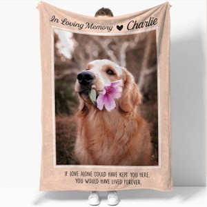 May include: A pink and beige memorial blanket with a photo of a golden retriever holding a pink flower in its mouth. The text "In Loving Memory Charlie" is at the top of the blanket and the text "If love alone could have kept you here, you would have lived forever." is at the bottom.