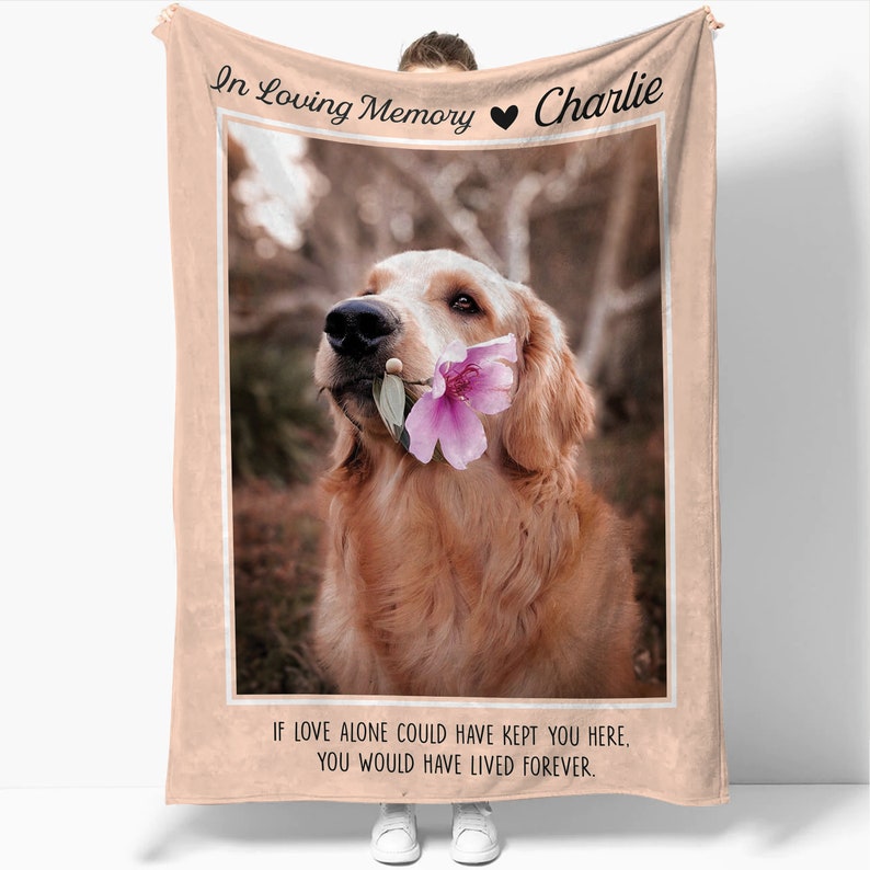 May include: A pink and beige memorial blanket with a photo of a golden retriever holding a pink flower in its mouth. The text "In Loving Memory Charlie" is at the top of the blanket and the text "If love alone could have kept you here, you would have lived forever." is at the bottom.