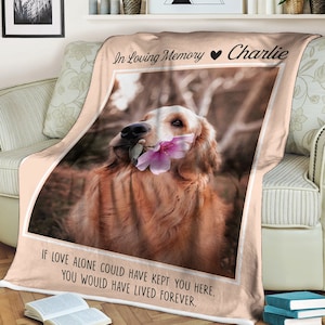 May include: A pink and white memorial blanket with a photo of a golden retriever holding a pink flower in its mouth. The text "In Loving Memory Charlie" is at the top of the blanket and the text "IF LOVE ALONE COULD HAVE KEPT YOU HERE, YOU WOULD HAVE LIVED FOREVER." is at the bottom.