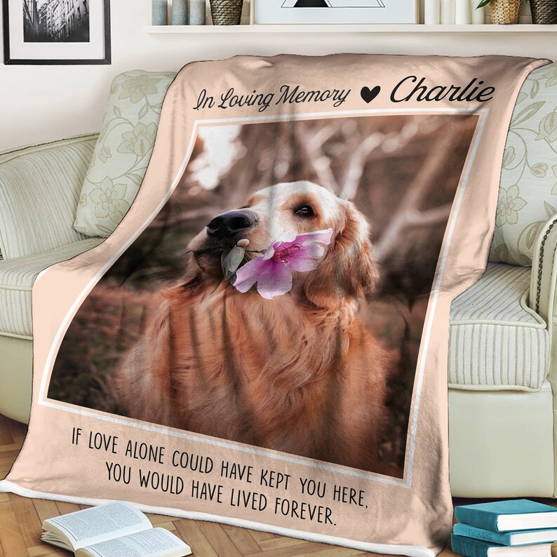 May include: A pink and white memorial blanket with a photo of a golden retriever holding a pink flower in its mouth. The text "In Loving Memory Charlie" is at the top of the blanket and the text "IF LOVE ALONE COULD HAVE KEPT YOU HERE, YOU WOULD HAVE LIVED FOREVER." is at the bottom.