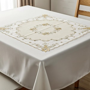 May include: A white tablecloth with a square, embroidered centerpiece. The centerpiece features a floral design in gold thread, with a scalloped edge. The tablecloth is set on a wooden table.