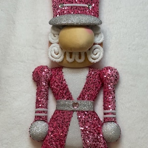 May include: A pink and silver decorative nutcracker figurine. The figurine has a pink sequined jacket, hat, and arms, with silver accents on the hat, belt, and arm balls. The face is a light tan color with white curly hair.
