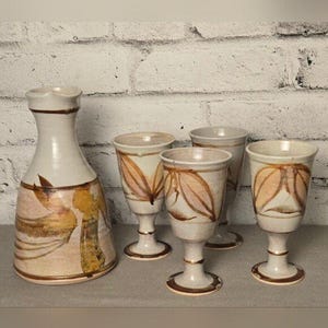 May include: A ceramic jug and four matching goblets. The jug is off-white with brown and gold abstract designs. The goblets have a similar design, with a brown leaf pattern. The set is on a grey surface against a white brick wall.