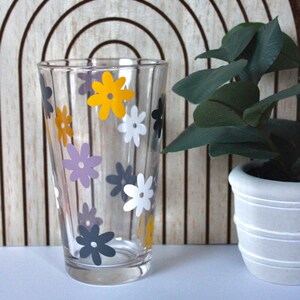 Puede incluir: Un vaso de vidrio transparente con un patrón floral. El vaso tiene flores amarillas, blancas y grises. El vaso está sobre una superficie blanca frente a un fondo de madera.