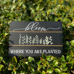 May include: Black wooden sign with white text that reads "bloom where you are planted". The sign has a row of green plant silhouettes.