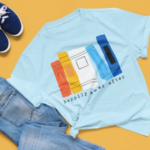 May include: Pale blue t-shirt with a graphic of stylised books in orange, yellow, white, light blue, and dark blue, with the words "happily ever after" printed below. The shirt is shown with blue jeans and trainers.