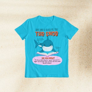 May include: A turquoise blue t-shirt with a white shark illustration holding a book. The text on the shirt reads "What kind of reader are you? The Snob" and "Did you know? The Great White Shark is 'great' because it's bigger and stronger than another fish also called the White Shark."