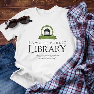 May include: White t-shirt featuring a green logo that reads "Pawnee Public Library" and the text "The worst group of people ever assembled in history."