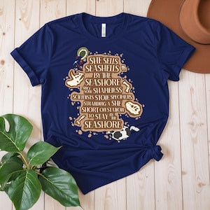 May include: A navy blue t-shirt featuring the phrase "SHE SELLS SEASHELLS BY THE SEASHORE" in a brown, vintage design. The design incorporates illustrations of seashells and marine elements. The t-shirt is knotted at the bottom.