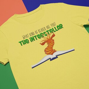 May include: Yellow t-shirt with a green graphic of a book with a red and orange creature on top. The text "What kind of reader are you? The interstellar" is printed on the shirt.