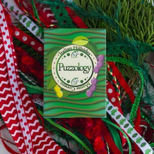 May include: A green and white game box with the text "Italian Hillsides Monte Corno Puzzology" on the front. The box is surrounded by red and white ribbon and green foliage.