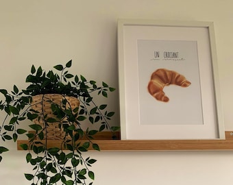 Könnte beinhalten: Ein weißer gerahmter Druck mit einer braunen Croissant-Illustration und dem Text "UN CROISSANT" in Schwarz. Der Druck hängt an einem Holzregal mit einer grünen Pflanze in einem Weidenkorb.