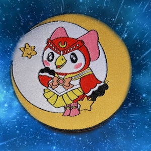 May include: A yellow and white embroidered patch featuring a cartoon owl wearing a red and white sailor outfit with a yellow star on the skirt. The owl is standing on a crescent moon with a yellow star in the background.