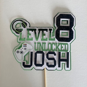 May include: Green and black cake topper with a video game controller, headphones, the number 8, and the text "Level 8 Unlocked Josh".