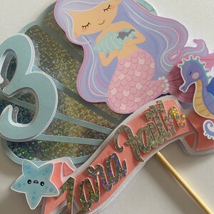May include: A mermaid cake topper with a purple and pink mermaid holding a seashell. The topper also includes a blue starfish, a seahorse, and a pink banner with the name "Zara Faith" in gold glitter letters.