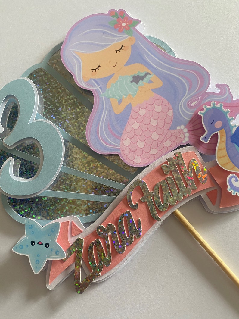 May include: A mermaid cake topper with a purple and pink mermaid holding a seashell. The topper also includes a blue starfish, a seahorse, and a pink banner with the name "Zara Faith" in gold glitter letters.