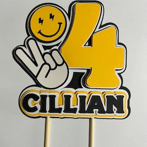 May include: Yellow and black cake topper with the number 4, a peace sign, a smiley face with lightning bolts, and the name "Cillian".