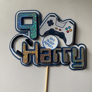 May include: A blue and silver cake topper with the number 9, the name Harry, a video game controller, and headphones. The controller has a white background with black buttons and a red and yellow accent. The text on the cake topper reads "Eat Sleep Repeat".