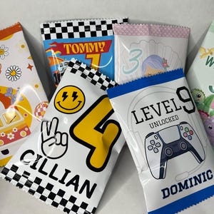 May include: Nine colorful party favor bags with different designs and names. The bags feature a variety of themes, including a car, a smiley face, a video game controller, a mermaid, and a dinosaur. The bags are all decorated with colorful patterns and text.