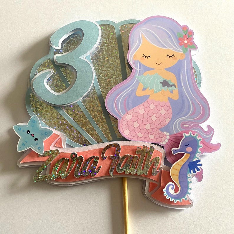 May include: A mermaid cake topper with a purple and pink mermaid holding a seashell. The topper also includes a blue number 3, a starfish, and a purple and blue seahorse. The topper is decorated with glitter and the words "Zara Faith".