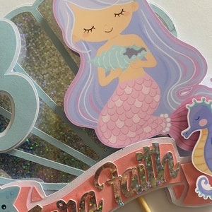 May include: A mermaid cake topper with a pink tail, purple hair, and a seashell in her hands. The topper is on a stick and has a pink banner with the name "Zara Faith" in silver glitter letters.