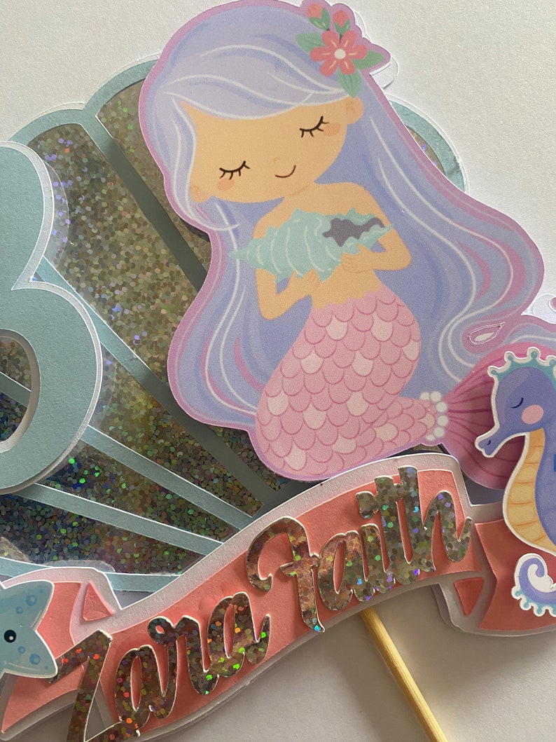 May include: A mermaid cake topper with a pink tail, purple hair, and a seashell in her hands. The topper is on a stick and has a pink banner with the name "Zara Faith" in silver glitter letters.