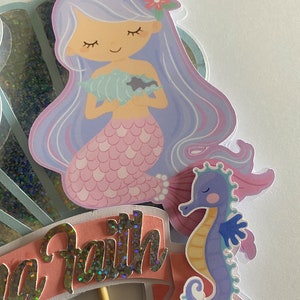 May include: A mermaid cake topper with a purple tail, a pink seashell, and a purple seahorse. The mermaid has long purple hair and a pink flower in her hair. The cake topper says "Sea Faith" in silver glitter letters.
