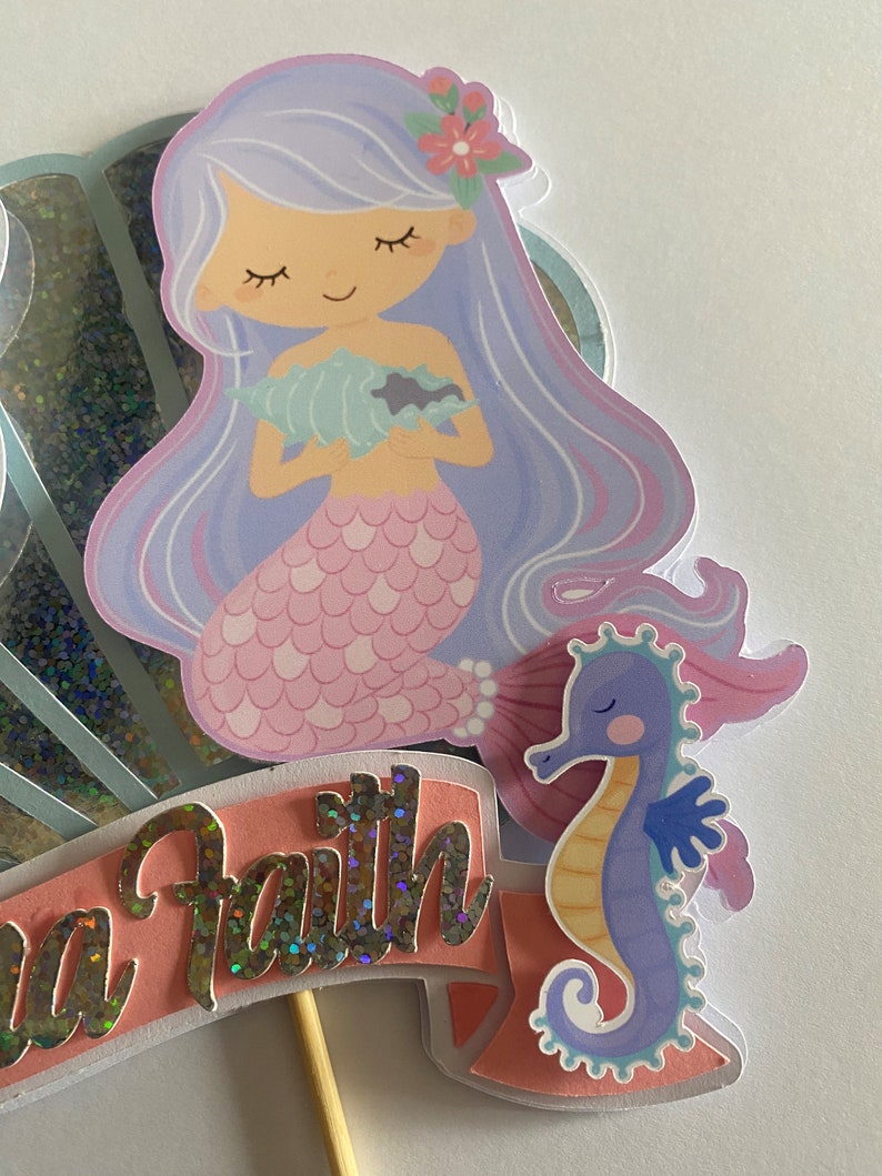 May include: A mermaid cake topper with a purple tail, a pink seashell, and a purple seahorse. The mermaid has long purple hair and a pink flower in her hair. The cake topper says "Sea Faith" in silver glitter letters.