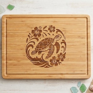 May include: A rectangular bamboo cutting board featuring a laser-engraved design of a sea turtle swimming amidst stylized ocean waves and hibiscus flowers. The board has a grooved perimeter channel and a natural light brown wood grain finish.