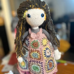 Könnte beinhalten: Häkelpuppe mit braunen Garnhaaren, einem rosa Kleid mit bunten Quadraten und blauen Augen.