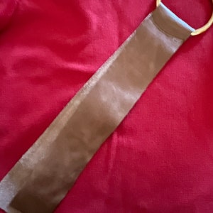 May include: A brown leather strap with a gold metal ring at one end. The strap is lying on a red fabric surface.