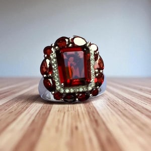 May include: A silver ring with a large, rectangular, deep red gemstone at its centre, encircled by smaller, round red stones and clear accent stones. The ring is set on a wooden surface.