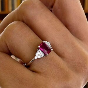 Puede incluir: Un anillo de plata con una llamativa gema roja rectangular como punto focal. El anillo est&aacute; adornado con gemas claras m&aacute;s peque&ntilde;as a ambos lados de la piedra principal, creando un dise&ntilde;o elegante y brillante. El anillo est&aacute; engarzado en una delgada banda de plata.