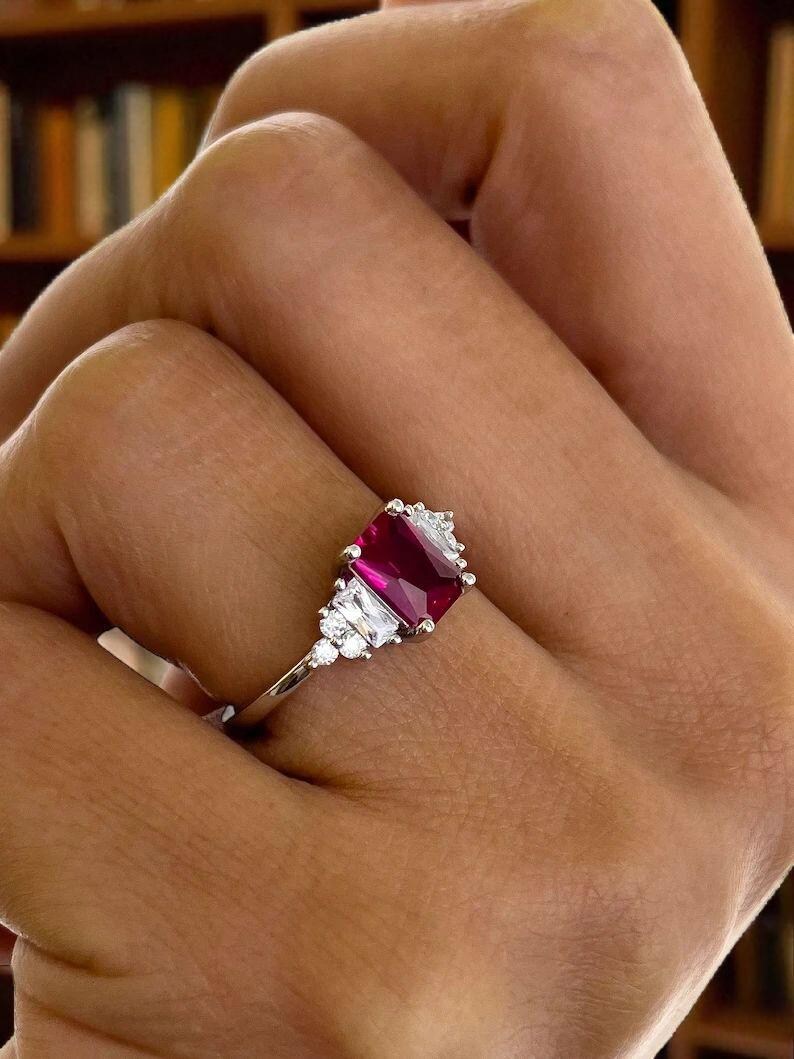 Puede incluir: Un anillo de plata con una llamativa gema roja rectangular como punto focal. El anillo est&aacute; adornado con gemas claras m&aacute;s peque&ntilde;as a ambos lados de la piedra principal, creando un dise&ntilde;o elegante y brillante. El anillo est&aacute; engarzado en una delgada banda de plata.