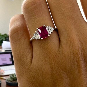 Puede incluir: Un anillo de plata con una gran gema rectangular de color rojo intenso como pieza central. A ambos lados de la piedra central, hay gemas m&aacute;s peque&ntilde;as, claras, rectangulares y redondas. El anillo se muestra en una mano.