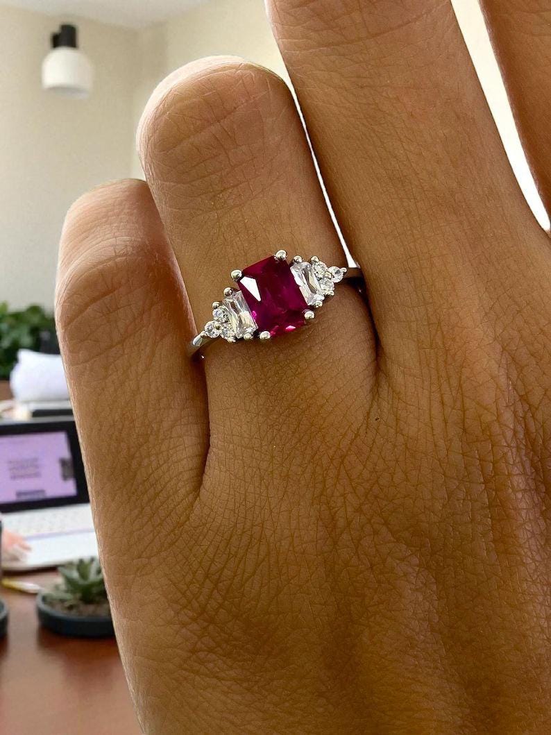 Puede incluir: Un anillo de plata con una gran gema rectangular de color rojo intenso como pieza central. A ambos lados de la piedra central, hay gemas m&aacute;s peque&ntilde;as, claras, rectangulares y redondas. El anillo se muestra en una mano.