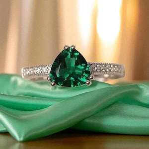 Op de afbeelding: Een zilveren ring met een grote, driehoekige, smaragdgroene edelsteen. De band is versierd met kleine, heldere edelstenen. De ring wordt gepresenteerd op groene satijnen stof.
