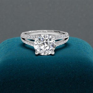 May include: A silver engagement ring with a large, round-cut diamond as the centerpiece. The band features a split shank design, with smaller diamonds set along the band. The ring is displayed on a teal surface.