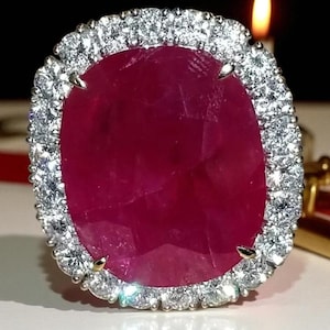 May include: A large, oval-cut ruby ring with a halo of small, round diamonds. The ruby is a deep red color, and the diamonds sparkle brightly. The ring is set in a silver-colored metal.