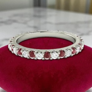 May include: A silver ring with alternating round-cut white and red gemstones. The ring is set on a red velvet surface, showcasing the sparkle of the gems. The ring's design features a continuous band of stones.