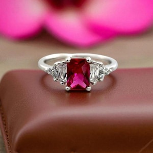 Könnte beinhalten: Ein silberner Ring mit einem großen, rechteckigen, leuchtend roten Edelstein im Mittelpunkt. Flankiert wird der zentrale Stein von zwei rechteckigen weißen Edelsteinen, mit kleineren weißen Steinen auf beiden Seiten. Der Ring liegt auf einer braunen Oberfläche, mit einem rosa Blumengrund.