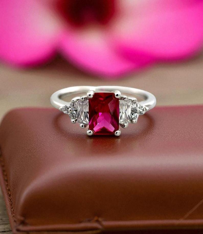 Puede incluir: Un anillo plateado con una gran gema roja rectangular vibrante como pieza central. A ambos lados de la piedra central hay dos gemas blancas rectangulares, con piedras blancas m&aacute;s peque&ntilde;as. El anillo est&aacute; sobre una superficie marr&oacute;n, con un fondo floral rosa.