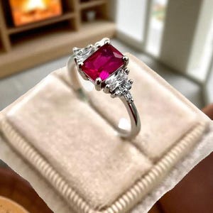Puede incluir: Un anillo plateado con una gran piedra central de color rub&iacute; cuadrado. El anillo est&aacute; flanqueado por piedras m&aacute;s peque&ntilde;as y transparentes a cada lado y est&aacute; sobre una superficie de terciopelo beige.